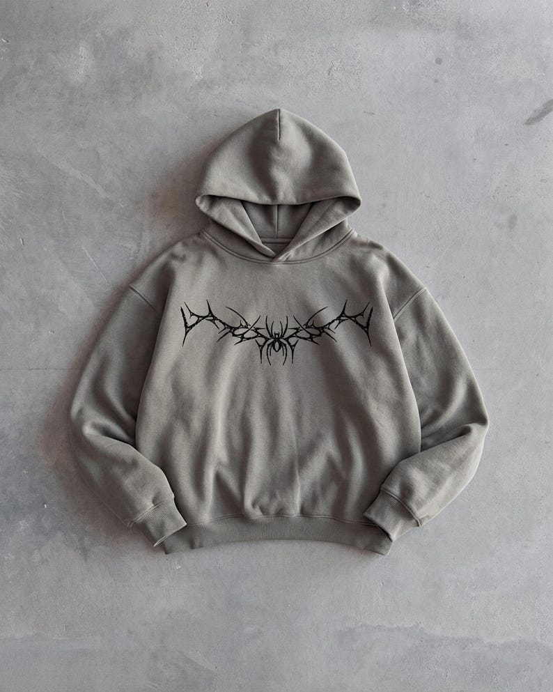 Puede incluir: Una sudadera con capucha gris con un dise&ntilde;o gr&aacute;fico negro en el pecho. El dise&ntilde;o parece ser un emblema estilizado y puntiagudo. La sudadera tiene capucha y mangas largas. La tela parece ser un material suave y de peso medio.