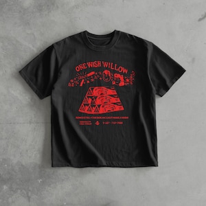 Puede incluir: Camiseta negra con texto y gráficos rojos. El diseño incluye las palabras "ONE WISH WILLOW" y una forma de pirámide. Debajo, el texto dice "REMOVE FRO...THE BOX AND JUST MAKE A WISH!" y un número de teléfono.