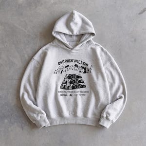 May include: A light gray hoodie with a hood and long sleeves. The front features a black graphic with the text "ONE WISH WILLOW" and the phrase "REMOVE PRO THE BOX AND JUST MAKE A WISH". The hoodie has a relaxed fit.