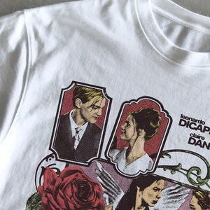 Pu&ograve; includere: Una t-shirt bianca con una stampa grafica di Leonardo DiCaprio e Claire Danes in ritratti incorniciati, accompagnata da una grande rosa rossa, una figura alata e il testo "leonardo DICAPRIO" e "claire DANES" in lettere nere.