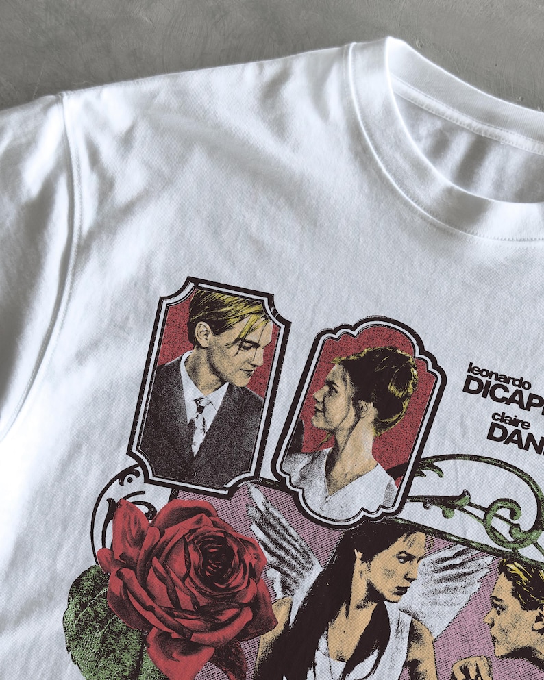 Pu&ograve; includere: Una t-shirt bianca con una stampa grafica di Leonardo DiCaprio e Claire Danes in ritratti incorniciati, accompagnata da una grande rosa rossa, una figura alata e il testo "leonardo DICAPRIO" e "claire DANES" in lettere nere.