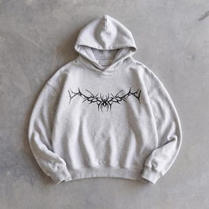 Puede incluir: Sudadera con capucha gris claro con un dise&ntilde;o gr&aacute;fico negro en el pecho. El dise&ntilde;o presenta un patr&oacute;n sim&eacute;trico y puntiagudo con un elemento similar a una ara&ntilde;a en el centro. La sudadera tiene capucha y mangas largas.