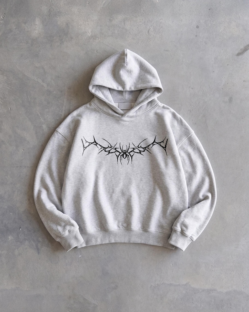 Puede incluir: Sudadera con capucha gris claro con un dise&ntilde;o gr&aacute;fico negro en el pecho. El dise&ntilde;o presenta un patr&oacute;n sim&eacute;trico y puntiagudo con un elemento similar a una ara&ntilde;a en el centro. La sudadera tiene capucha y mangas largas.