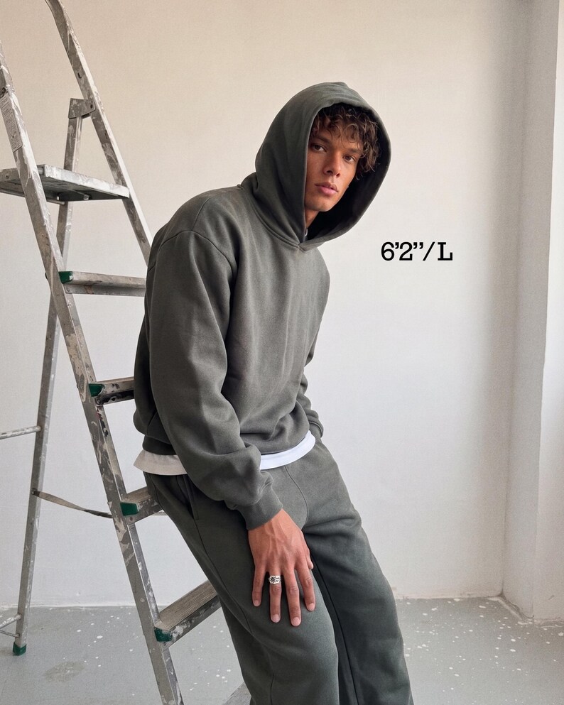 Puede incluir: Un conjunto de sudadera con capucha y pantal&oacute;n de ch&aacute;ndal verde oscuro. El modelo lleva una camiseta interior blanca. El texto de la imagen dice "6'2"/L". La persona est&aacute; apoyada en una escalera de metal.