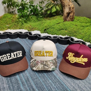 May include: Three baseball caps with curved brims. One is black with "GREATER" in white, another is cream with "GREATER" in gold, and the third is burgundy with "Kingdom" in gold. All have brown brims.