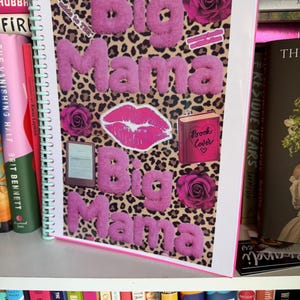 Op de afbeelding: Een spiraalgebonden notitieboek met een omslag met de woorden "Big Mama" in pluizige roze letters. De achtergrond is een luipaardprint. De omslag bevat ook roze rozen, een lipafdruk en een boekcover-afbeelding.