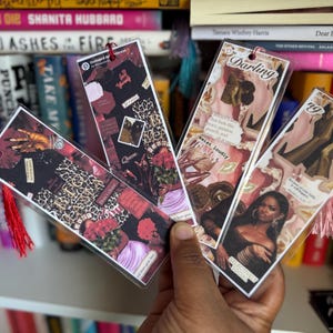 May include: Four decorative bookmarks with red tassels are held in front of a bookshelf. The bookmarks feature collages with images of women, leopard print, and text including "Darling" and "exist loudly".