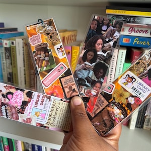 May include: A hand holds several clear, plastic bookmarks with colorful designs. The bookmarks feature images of people, flowers, and text like "Book Boyfriend" and "Library Card." Tassels hang from the top of some bookmarks. Bookshelves filled with books are in the background.