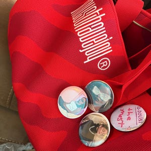 May include: A red tote bag with a striped pattern and the word "Notorious" in white. Four round badges with images and text are on the bag. One badge says "the most" and another says "to-dolist".