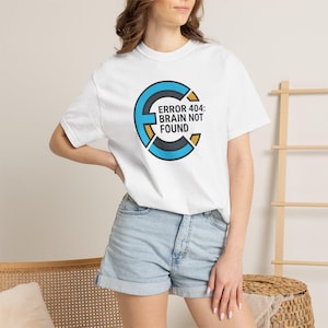 Op de afbeelding: Wit T-shirt met de tekst "ERROR 404: BRAIN NOT FOUND". Het ontwerp toont een gestileerde letter "E" in blauw, zwart en goud. Het model draagt een spijkerbroekshort.