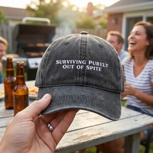 May include: A charcoal gray baseball cap with a distressed, washed-out finish. The front panel features white embroidered text that reads, "SURVIVING PURELY OUT OF SPITE." The cap has a curved brim and visible stitching along the crown and visor.