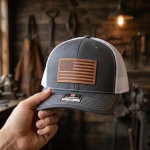 May include: A gray and white trucker hat with a leather American flag patch. The hat has a mesh back and a curved brim. The patch is rectangular and features the flag's stripes and stars. The hat is being held up by a hand.