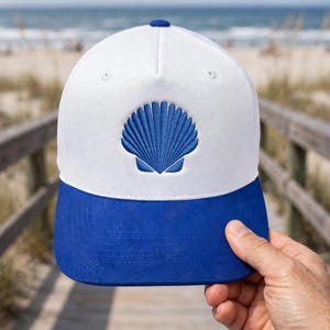 May include: A white baseball cap featuring a blue embroidered scallop shell design on the front panel. The cap has a solid blue brim and a matching blue button at the top center, constructed with a structured crown and stitched eyelets for ventilation.