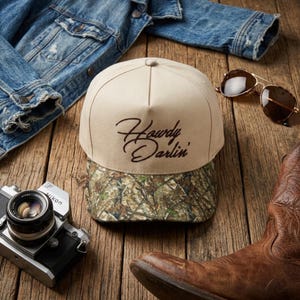 May include: A beige baseball cap with a structured crown and a camouflage print brim. The front of the cap features the text Howdy Darlin' embroidered in dark brown script. The cap is displayed alongside a denim jacket, sunglasses, a camera, and a boot.