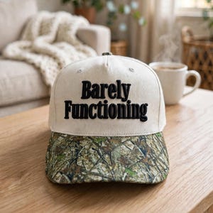 May include: A baseball cap with a cream-colored crown and a camouflage-patterned brim. The front of the crown features the text "Barely Functioning" embroidered in bold, black, raised lettering. The cap has a structured five-panel design.