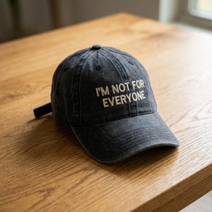 May include: A dark gray baseball cap with the embroidered text "I'M NOT FOR EVERYONE" in white. The cap is made of a worn-looking fabric and has a curved brim and an adjustable strap.