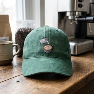 May include: A forest green baseball cap featuring an embroidered graphic of a silver pitcher pouring steamed milk into a brown coffee cup to create latte art. The cap has a structured crown, a curved brim, and a classic six-panel design.