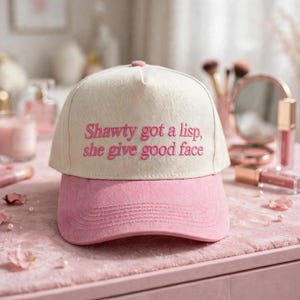 May include: A cream-colored baseball cap with a pink brim and pink embroidered text that reads, "Shawty got a lisp, she give good face." The cap features a structured front panel and a button on the top center, showcasing a two-tone design.