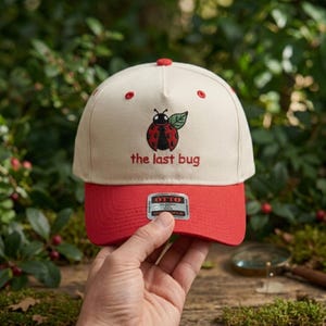 May include: A cream-colored baseball cap with a red brim and red eyelets. The front features an embroidered red and black ladybug with a green leaf and the red text "the last bug" underneath. An Otto brand sticker is centered on the brim.