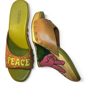 Op de afbeelding: Een paar beige leren sleehak sandalen met olijfgroene binnenzolen en houten zolen. De voorste band bevat het woord PEACE in gele en groene letters met kleine stervormige uitsparingen. De zijkant van de hak toont een roze peace-teken handafbeelding.