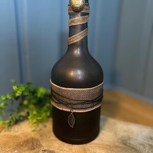 Op de afbeelding: Een decoratieve zwarte fles met gouden en zwarte accenten. De fles is omwikkeld met gouden touw en een zwart-beige band. Een kleine bladbedel hangt aan de band. De fles is afgewerkt met een gouden knop.