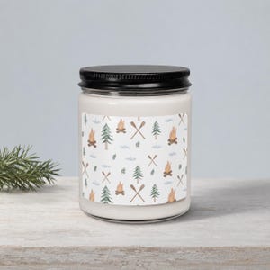 May include: A cylindrical glass candle jar with a black screw-top lid. The label features a repeating pattern of watercolor illustrations including orange campfires, green pine trees, crossed brown wooden oars, and small blue water ripples.