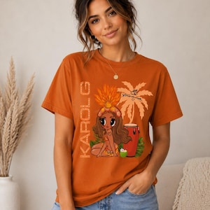 May include: A burnt orange short-sleeve t-shirt featuring a graphic print of a cartoon character wearing a feathered headdress, a palm tree, a conga drum, and the text KAROL G written vertically in a light-colored, dotted font style.