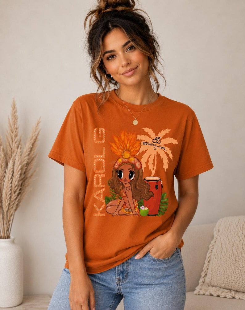 May include: A burnt orange short-sleeve t-shirt featuring a graphic print of a cartoon character wearing a feathered headdress, a palm tree, a conga drum, and the text KAROL G written vertically in a light-colored, dotted font style.