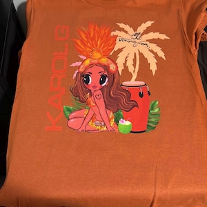 May include: A burnt orange t-shirt featuring a graphic print of a cartoon character with long brown hair, a feathered headpiece, and a heart tattoo. The design includes the text KAROL G, a palm tree, a red conga drum, a coconut drink, and green leaves.