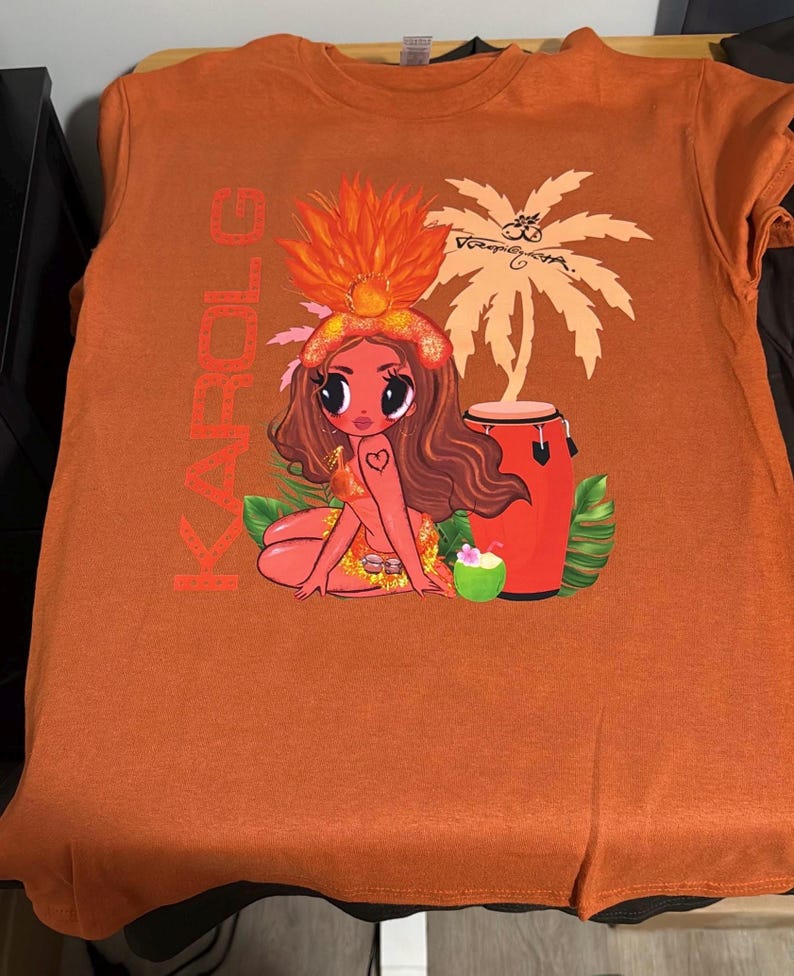 May include: A burnt orange t-shirt featuring a graphic print of a cartoon character with long brown hair, a feathered headpiece, and a heart tattoo. The design includes the text KAROL G, a palm tree, a red conga drum, a coconut drink, and green leaves.