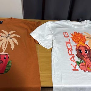 May include: Two short-sleeved t-shirts, one in burnt orange and one in white, each featuring a graphic print of a cartoon character with long brown hair, a feathered headdress, and a red conga drum, with the text KAROL G printed vertically.