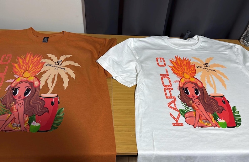 May include: Two short-sleeved t-shirts, one in burnt orange and one in white, each featuring a graphic print of a cartoon character with long brown hair, a feathered headdress, and a red conga drum, with the text KAROL G printed vertically.