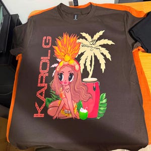 May include: A dark brown short-sleeve t-shirt featuring a graphic print of a cartoon character with long hair, a large orange headdress, and a bikini. The design includes the text KAROL G, a palm tree, a red conga drum, and a green coconut.