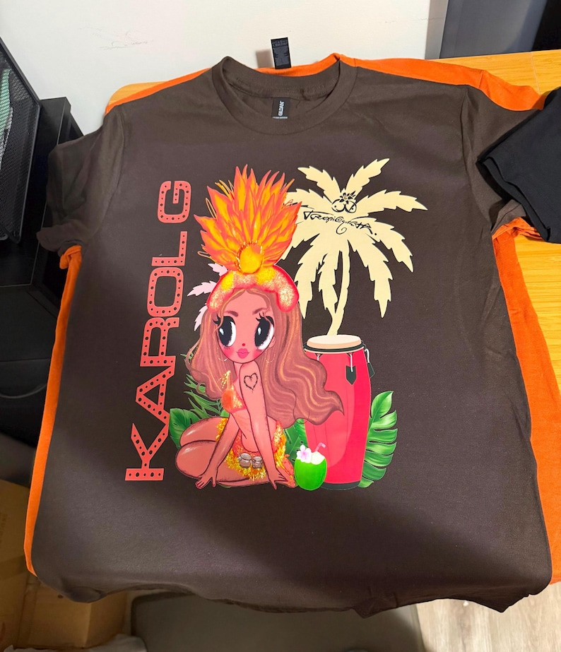 May include: A dark brown short-sleeve t-shirt featuring a graphic print of a cartoon character with long hair, a large orange headdress, and a bikini. The design includes the text KAROL G, a palm tree, a red conga drum, and a green coconut.
