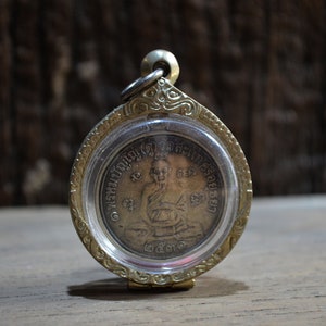 May include: A round, gold-colored pendant with a clear plastic cover. The pendant features a detailed image of a seated figure and text in a foreign language. The pendant is suspended from a small gold ring.