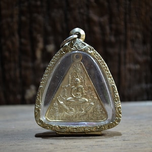 Puede incluir: Un colgante dorado con una imagen detallada de una figura sentada dentro de un marco triangular. El colgante está encerrado en una cubierta transparente y tiene un borde ornamentado. El fondo es una superficie de madera borrosa.