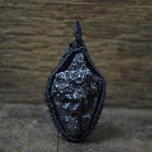 Può includere: Un ciondolo scuro di forma irregolare con una superficie ruvida e strutturata. Il ciondolo è racchiuso in un cordoncino nero intrecciato, creando una forma a goccia. Lo sfondo è una superficie di legno rustico.