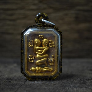 Puede incluir: Colgante rectangular dorado con cubierta transparente, que presenta un relieve de dos figuras abrazadas con un corazón encima. El colgante tiene un borde decorativo y un pequeño lazo para colgar. El fondo es una superficie oscura y texturizada.