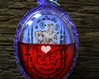 May include: A red and purple oval-shaped pendant with a white heart and a drawing of a man and a woman inside. The pendant has Thai writing around the edge.