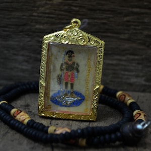 Può includere: Un ciondolo d'oro con un fronte di vetro che presenta un'immagine dipinta di una persona che tiene una lancia. Il ciondolo è su una collana di perline nere con perline di legno.