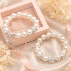 Puede incluir: Dos pulseras de perlas con detalles de pedrería. Una pulsera presenta un lazo y pedrería colgante, mientras que la otra tiene un centro de pedrería en forma de corazón. Las pulseras se exhiben sobre una superficie satinada.