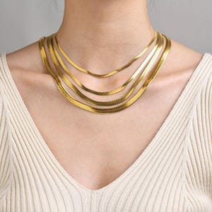 May include: A collection of gold-colored herringbone chain necklaces of varying lengths. The necklaces are layered and worn around the neck, creating a stylish and elegant look. The chains have a flat, smooth surface.