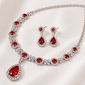 Puede incluir: Un conjunto de collar y pendientes plateados con piedras rojas en forma de lágrima. El collar tiene un colgante y está adornado con piedras rojas más pequeñas y cristales transparentes. Los pendientes reflejan el diseño.