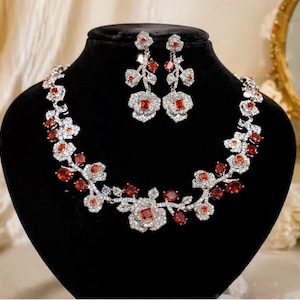 Puede incluir: Un conjunto de collar y pendientes exhibido en un maniquí negro. La joyería presenta un diseño floral con piedras preciosas claras y rojas. El collar se curva alrededor del cuello y los pendientes cuelgan.
