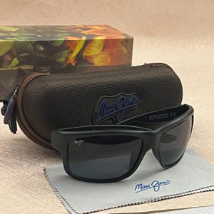 Może przedstawiać: Czarne okulary przeciwsłoneczne Maui Jim z ciemnymi soczewkami, posiadające logo marki na oprawce i soczewce. Okulary umieszczono przed brązowym, teksturowanym twardym etui z niebieskim logo oraz jasnoszarą ściereczką z mikrofibry do czyszczenia.
