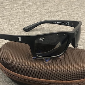 Puede incluir: Gafas de sol negras con lentes oscuras, sobre un estuche marr&oacute;n. Las gafas tienen un acabado mate y el texto "Manchester United" es visible en la patilla. El estuche tiene cremallera y una superficie texturizada.