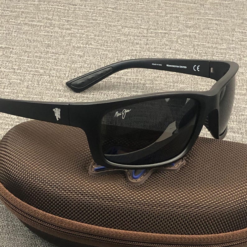 Puede incluir: Gafas de sol negras con lentes oscuras, sobre un estuche marr&oacute;n. Las gafas tienen un acabado mate y el texto "Manchester United" es visible en la patilla. El estuche tiene cremallera y una superficie texturizada.