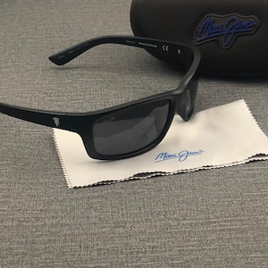 Puede incluir: Gafas de sol negras con lentes oscuras y un estuche a juego. Las gafas de sol descansan sobre un pa&ntilde;o de limpieza blanco con el nombre de la marca "Maui Jim" impreso en azul. El estuche tiene un logotipo azul.