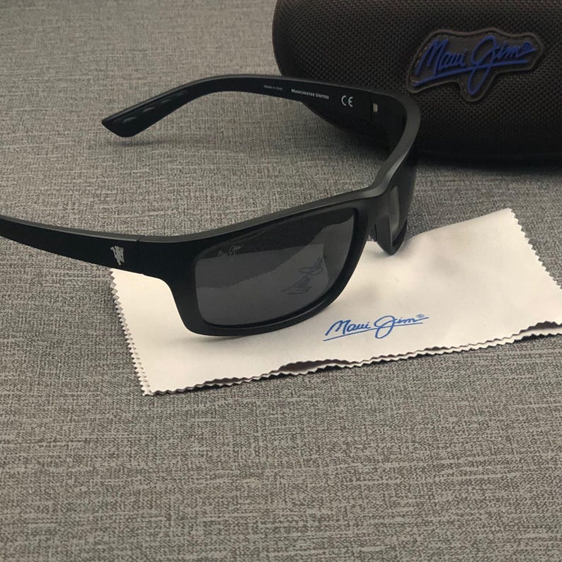 Puede incluir: Gafas de sol negras con lentes oscuras y un estuche a juego. Las gafas de sol descansan sobre un pa&ntilde;o de limpieza blanco con el nombre de la marca "Maui Jim" impreso en azul. El estuche tiene un logotipo azul.