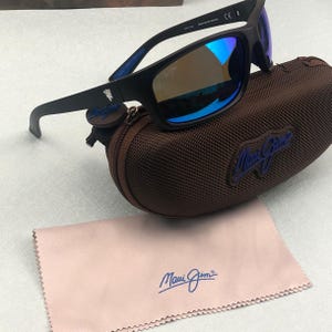 May include: Black sunglasses with blue mirrored lenses rest on a brown case. The sunglasses have a matte black frame and the brand name is visible on the case and cleaning cloth. A light pink cleaning cloth is also included.